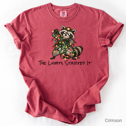 Headlamp-wearing raccoon tangled in colorful Christmas lights on a holiday graphic T-shirt